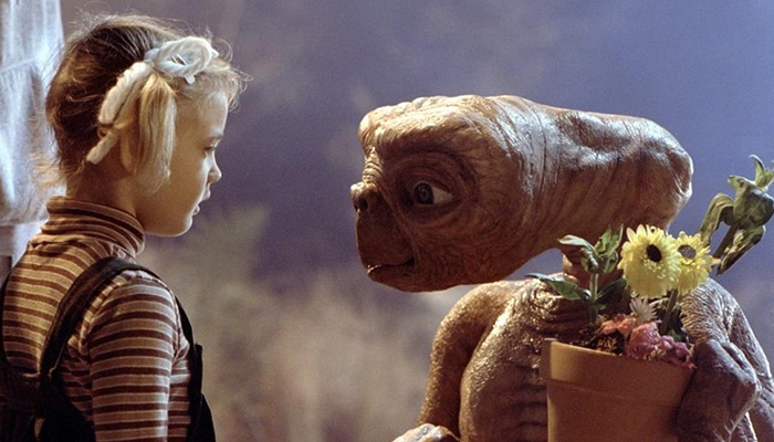 A young girl in ponytails (Drew Barrymore) looks at an alien holding a potted flower