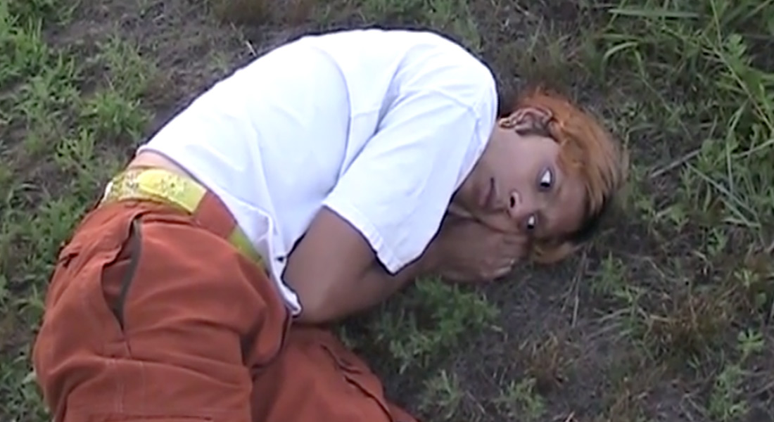 In this image, a person lays slightly curled up and wears a white t-shirt and orange pants.