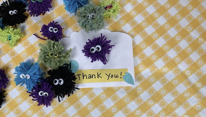 Pom pom creatures with googly eyes on a yellow checkered tablecloth and a sign that reads "thank you"
