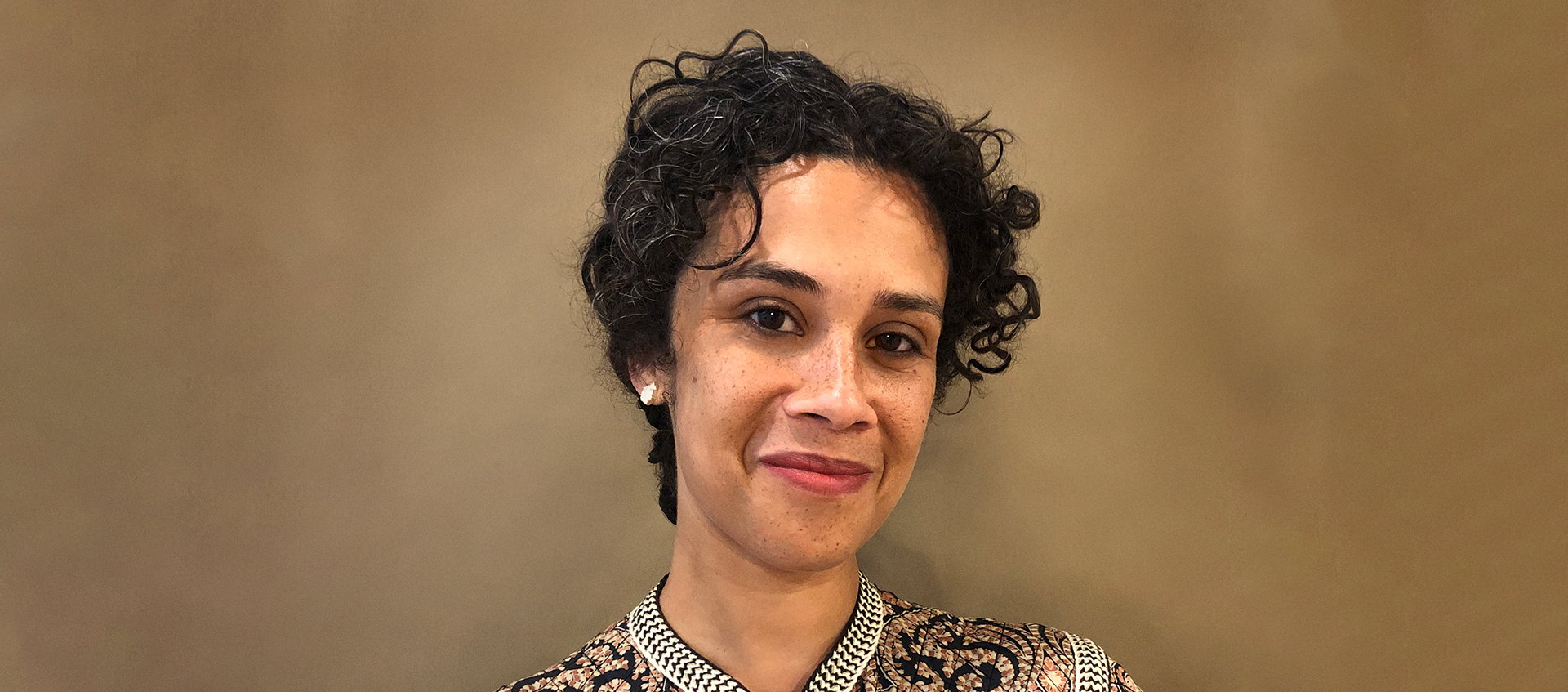 A photo of Jonna Twigg from the neck up. Jonna is in front of a brown background and wears earrings and has black hair.