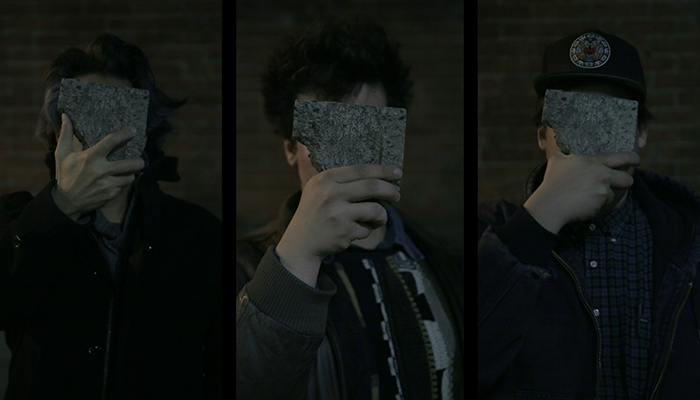 A photo of three members of New Red Order, they are all covering their faces with a piece of stone