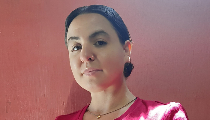 A photo of Tala Kanani in front of a reddish background. Tala has black hair and is wearing a red shirt, a gold necklace, and a nose ring.
