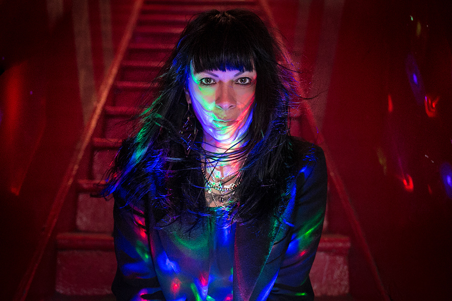 A color portrait of filmmaker Jennifer Reeder sitting in a dark stairwell lit by miniature color spotlights