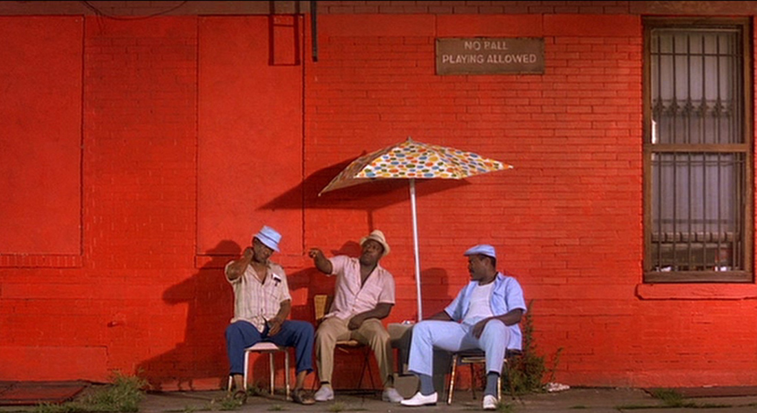Three Black men sit near a polka-dot umbrella against a red brick wall with a sign that reads “No Ball Playing Allowed” 