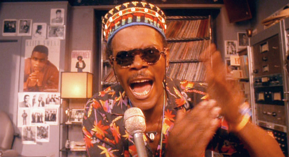 A man in round sunglasses and brightly patterned shirt and hat seems to shout at a microphone in a radio station