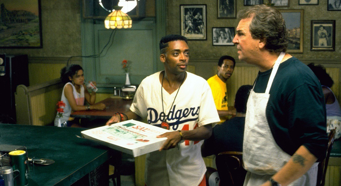 A young man in a Dodgers jersey holding a pizza box looks at an older man wearing an apron