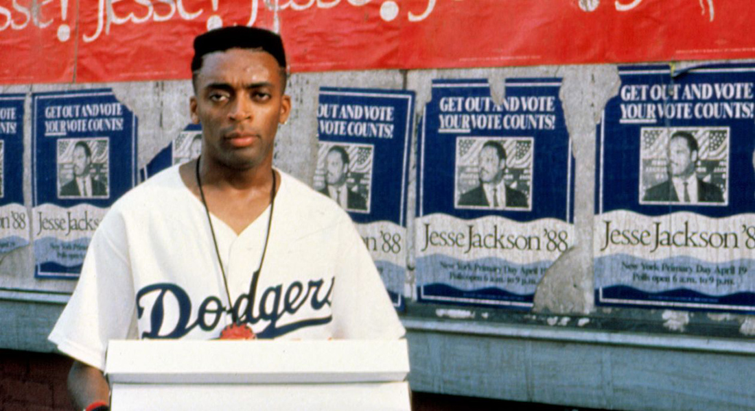 A young man in a Dodgers jersey holding pizza boxes looks directly at the camera in front of a wall of 1988 Jesse Jackson campaign posters that read "Get Out and Vote / Your Vote Counts!"