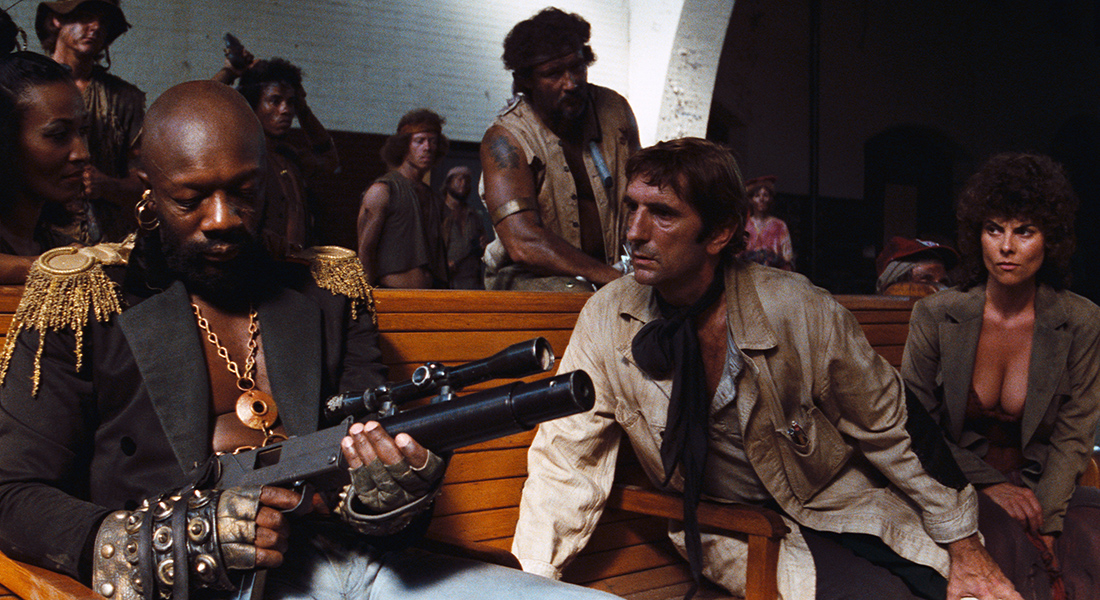 A seated, shirtless bald man in an antique jacket (Isaac Hayes) handles a modified pistol while two people on the same bench look on