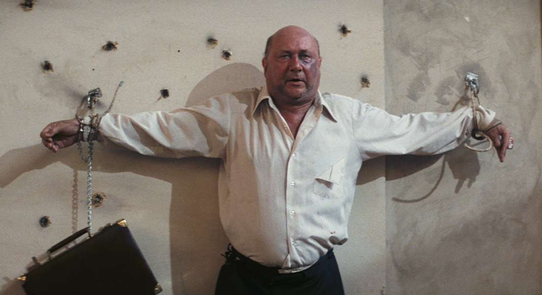 A man in a while shirt and a briefcase handcuffed to his left hand is roped to a wall just beneath a series of bullet holes