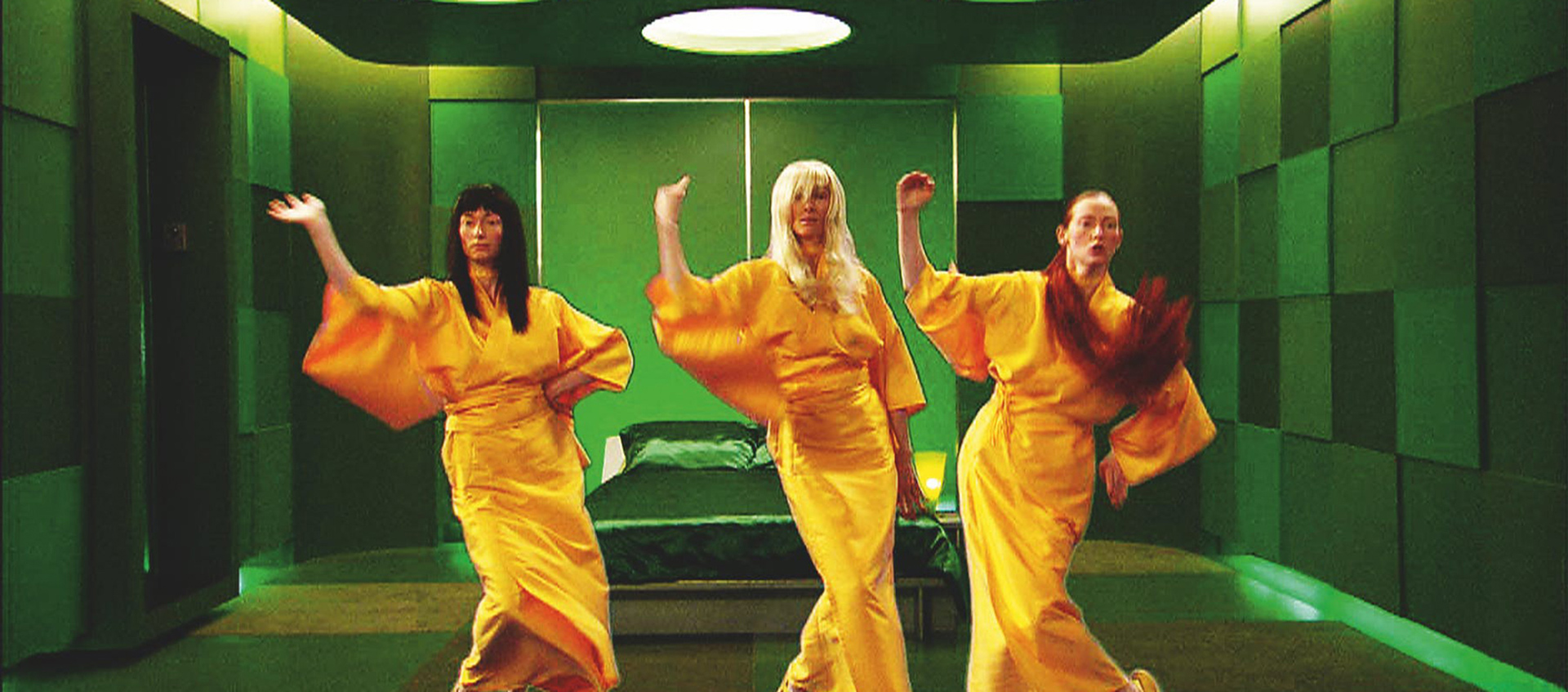 An image of three people all dressed in yellow outfits standing in an all green room. Each person has their right arm extended into the air.