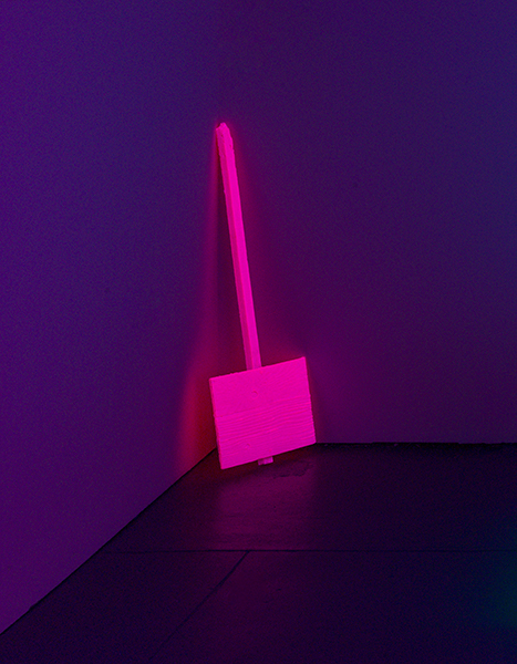 What appears to be a wooden sign on a post covered in flourescent pink paint stands on its head in the corner of a dark room with purple walls