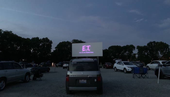 Cars in front of a drive-in screen with the opening credit for E.T.: the Extra-terrestrial