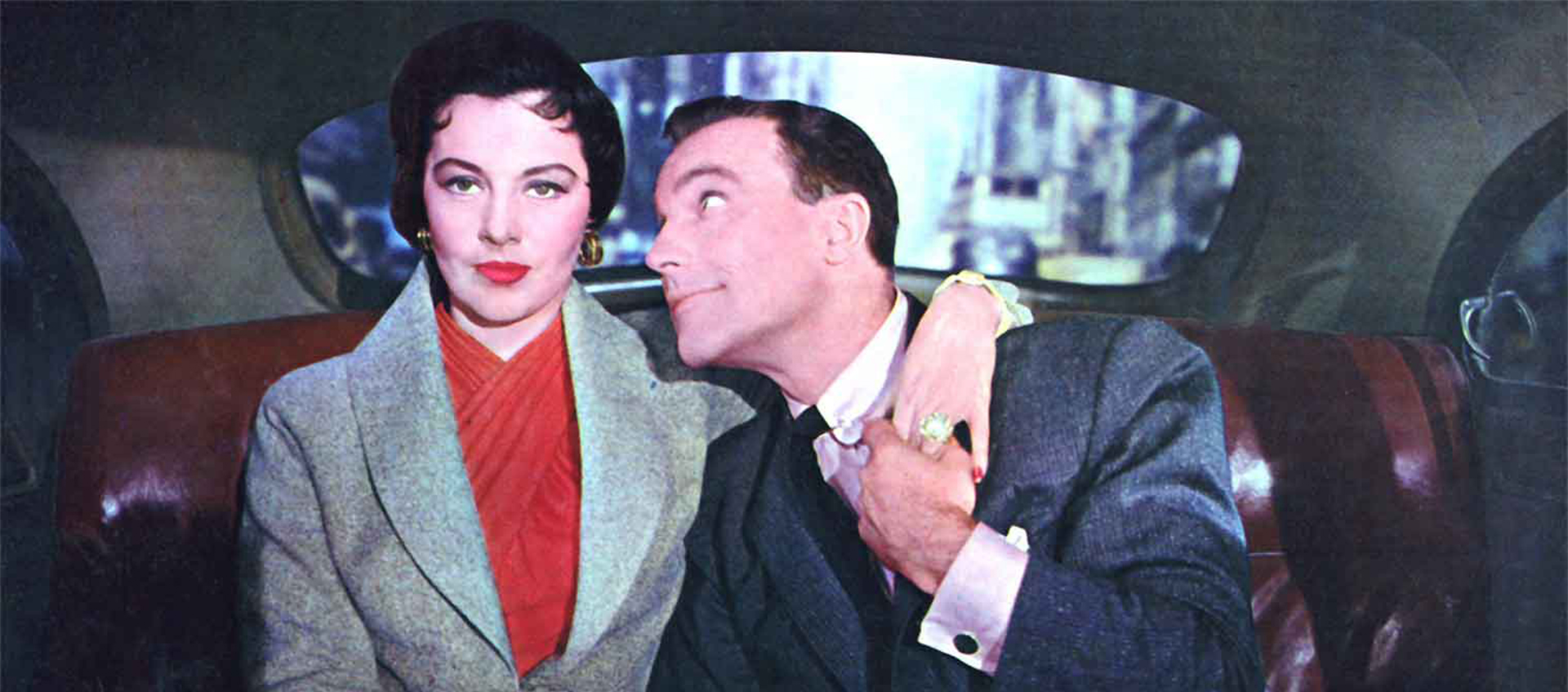 A couple sits in the backseat of a car. A woman with black hair pulled back in a gray jacket and red shirt has her arm around the shoulder of a man in a gray suit who looks longingly at her.