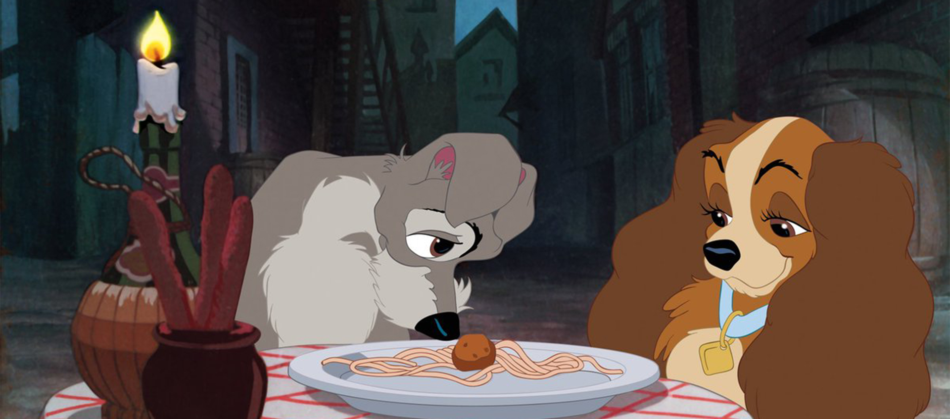 Two dogs seated at a table in front of a plate of spaghetti. On the left, a mutt pushes a meatball toward the cocker spaniel on the right with his nose.