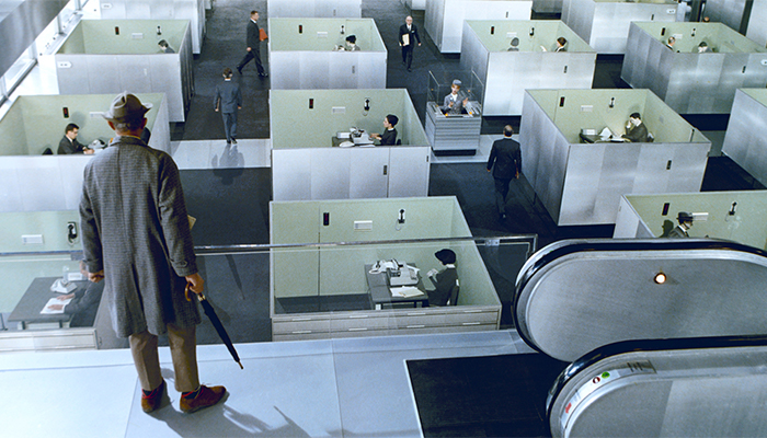 A person in a raincoat and hat looks over an office floor made up of dozens of cubicles. He holds an umbrella and stands on a level above the floor.