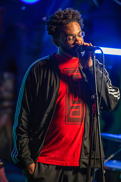 A vocalist in a black track jacket and red shirt sings in front of a microphone