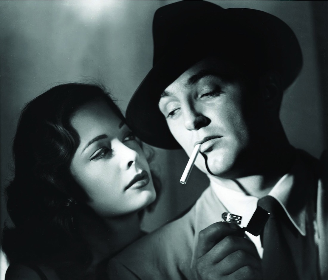 Black and white image of a woman on the left with dark hair looking at a man on the right who wears a hat and smokes a cigarette