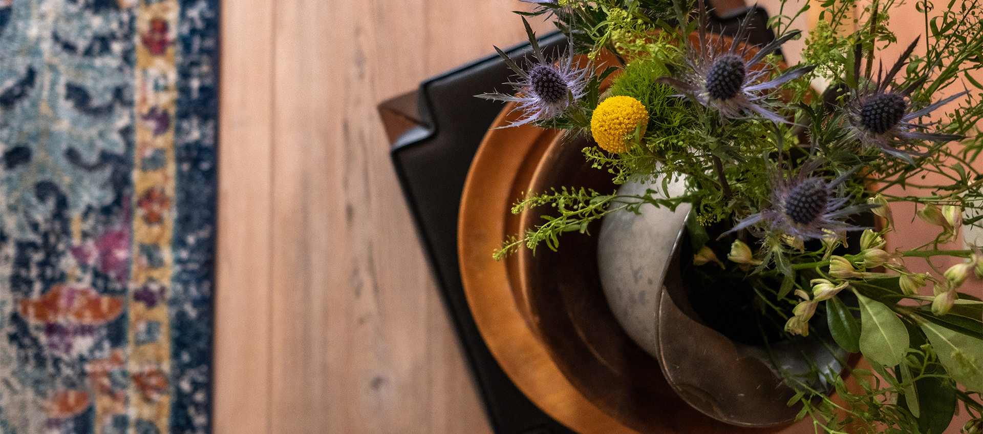 A floral arrangement of yellow craspedia and purple sea holly sits on a copper-colored tray on a small black chair as seen from above