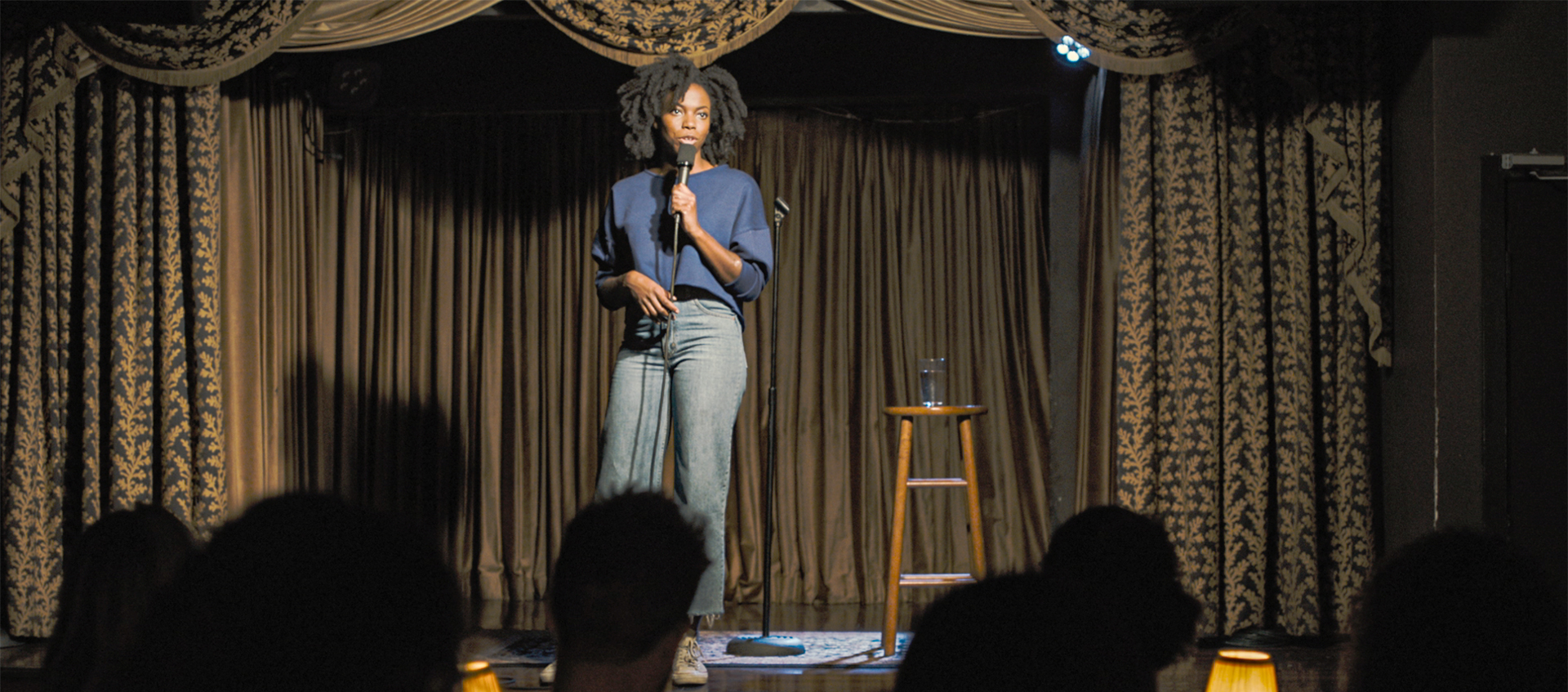 A woman on stage doing stand-up comedy. She stands in front of a stool and holds a microphone as she addresses the audiences. She wears a blue shirt and blue jeans
