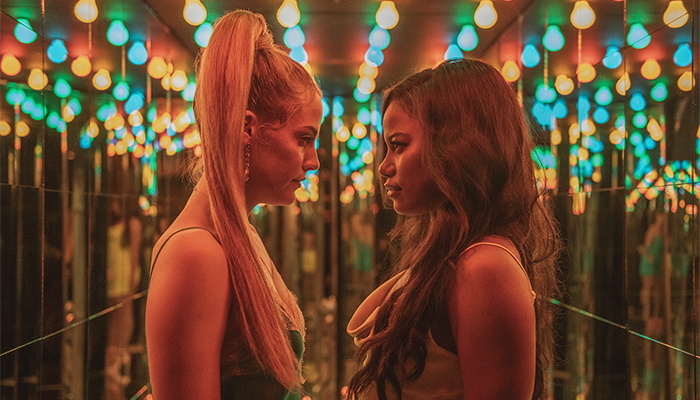 Two women stand in front of a mirrored surface and beneath rows of multicolored lights. The woman on the left has long blond hair in a ponytail and the woman on the right wears a pink top. They are both staring each other down.
