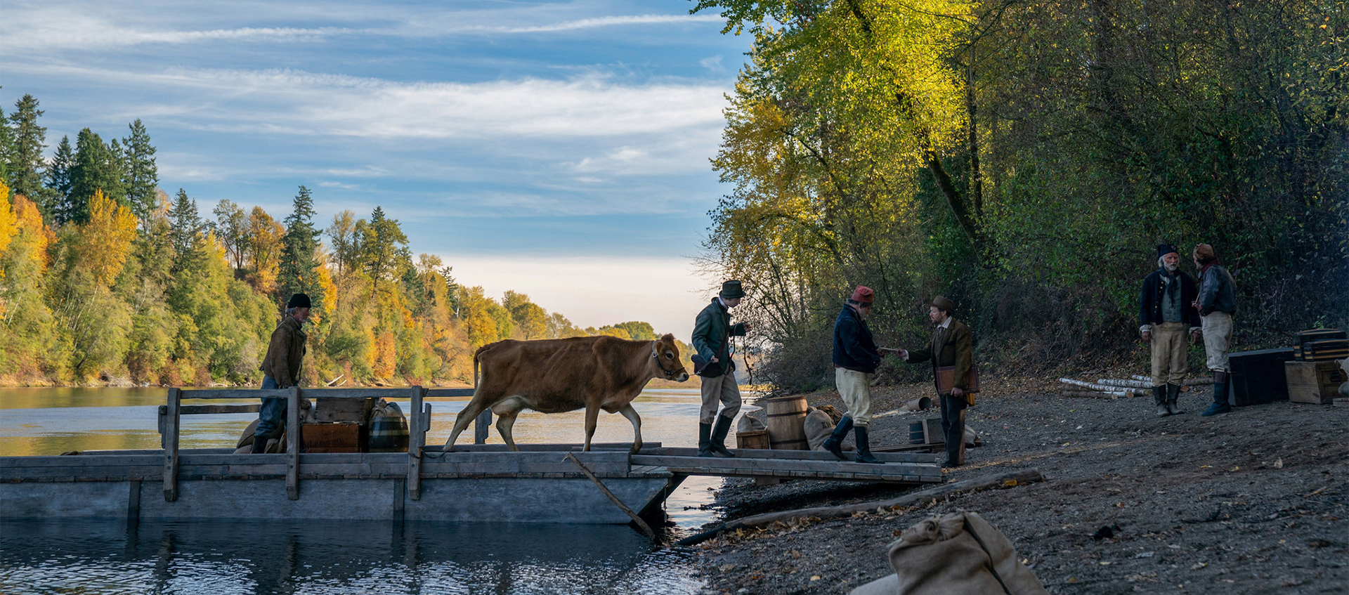 Three people cross a river by bridge. In between them a cow pulls suitcases and luggage. They approach the show where three other people wait for them.