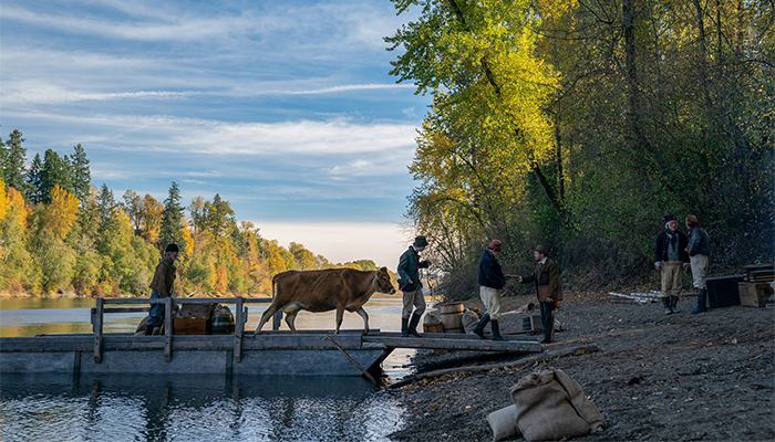 Three people cross a river by bridge. In between them a cow pulls suitcases and luggage. They approach the show where three other people wait for them.