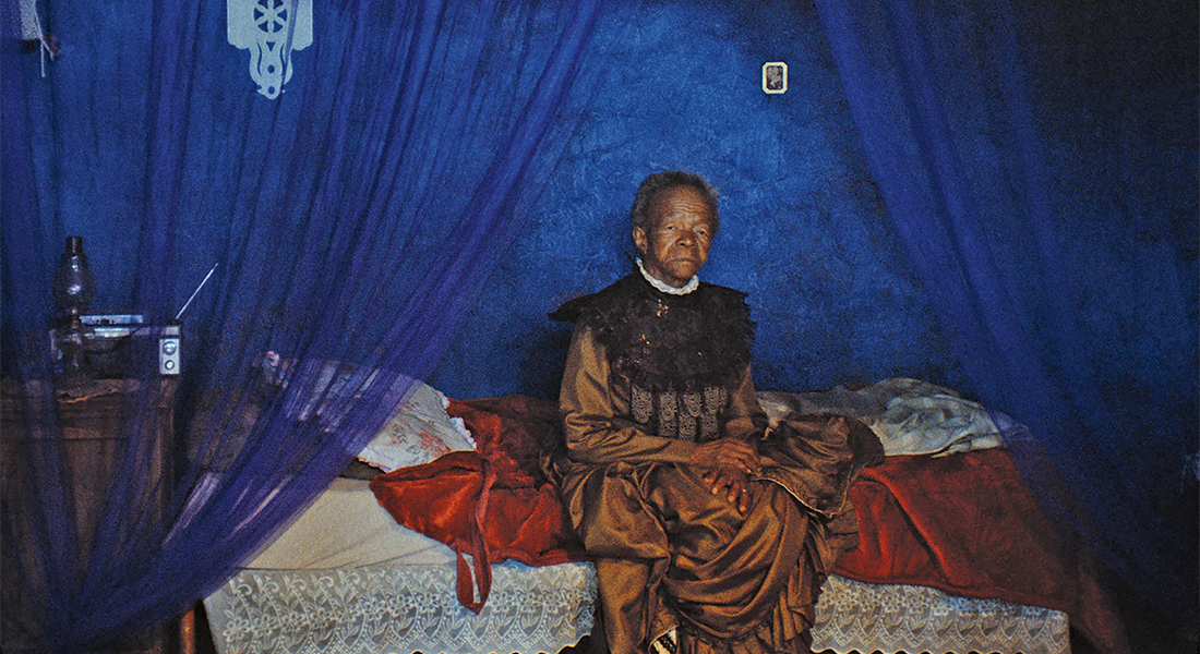 A person in a shiny brown gown sits on a bed in front of a bright blue wall. A radio is next to the bed.