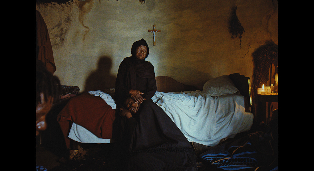A person in a dark robe with a hood sits with their hands crossed on their lap on a messy bed in front a light brown colored wall on which hangs a small crucifix.