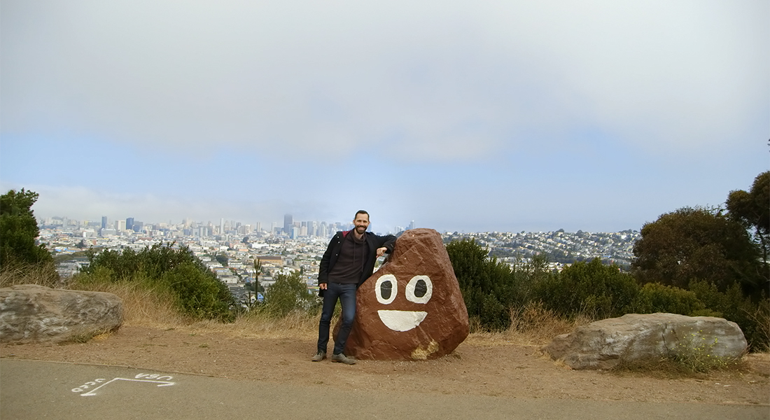 A person stands next to a rock painted to look like the poop emoji, which is a brown object with eyes 