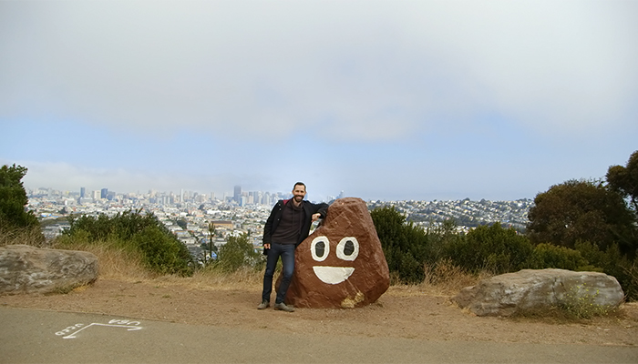A person stands next to a rock painted to look like the poop emoji, which is a brown object with eyes 