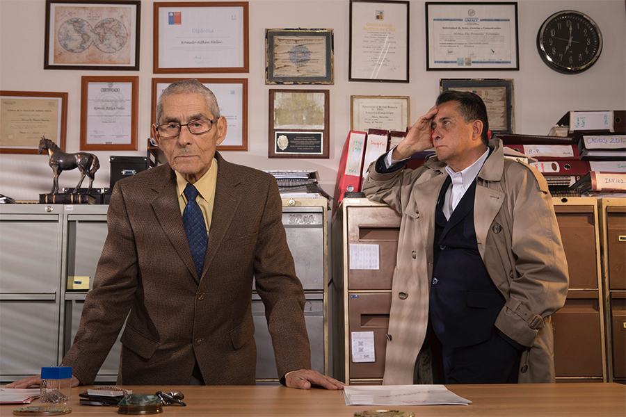 Two men, one in a brown suit and wearing glasses and the other in a trenchcoat with his hand resting on file cabinet stand in a cluttered office in front of file cabinets and a wall of photos and documents.