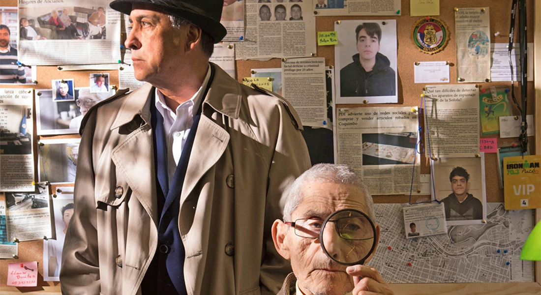 Two men in an office, one seated at a desk and holding a magnifying glass up to his eye and the other standing and in a trench coat and wearing a fedora and he looks out the window,