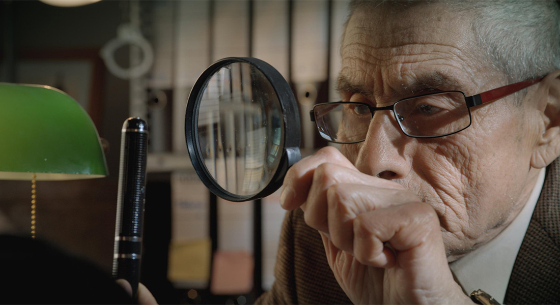 A close-up image of a man wearing eyeglasses and holding a magnifying glass up to his eyes.
