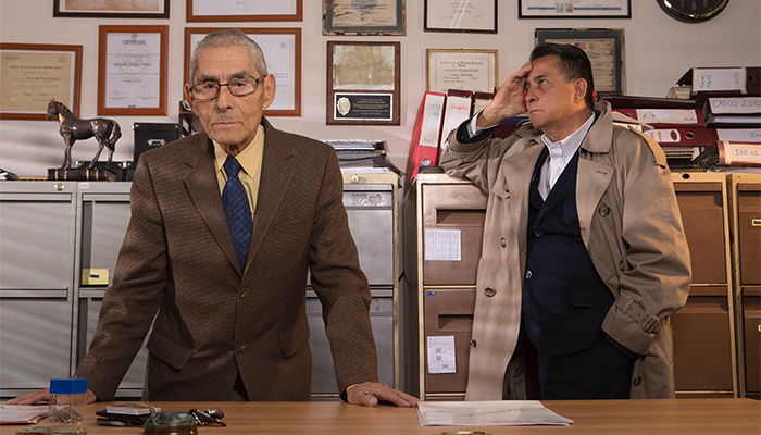 Two men, one in a brown suit and wearing glasses and the other in a trenchcoat with his hand resting on file cabinet stand in a cluttered office in front of file cabinets and a wall of photos and documents.
