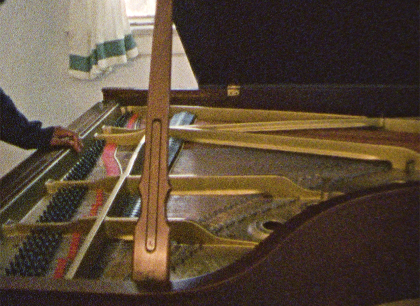 A piano with the lid open, exposing the strings and hammers inside. A person's hand rest on the edge of the piano.