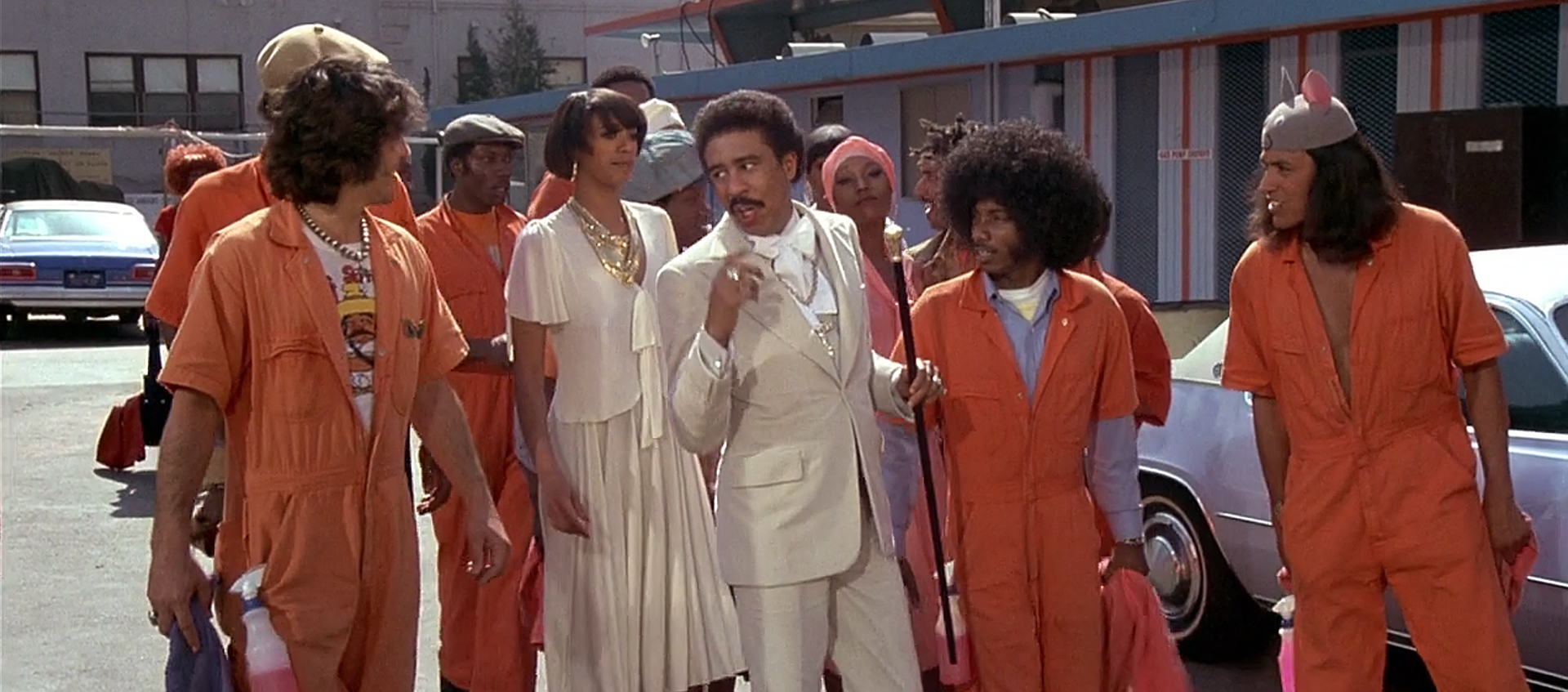 A group of nine people standing in front of a car. Eight of the people wear orange worksuits, while the two people in the center wear shiny white suits