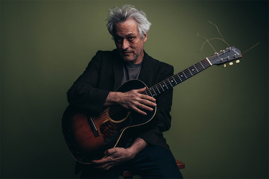 Marc Ribot, a man with gray hair and a black jacket holds a guitar