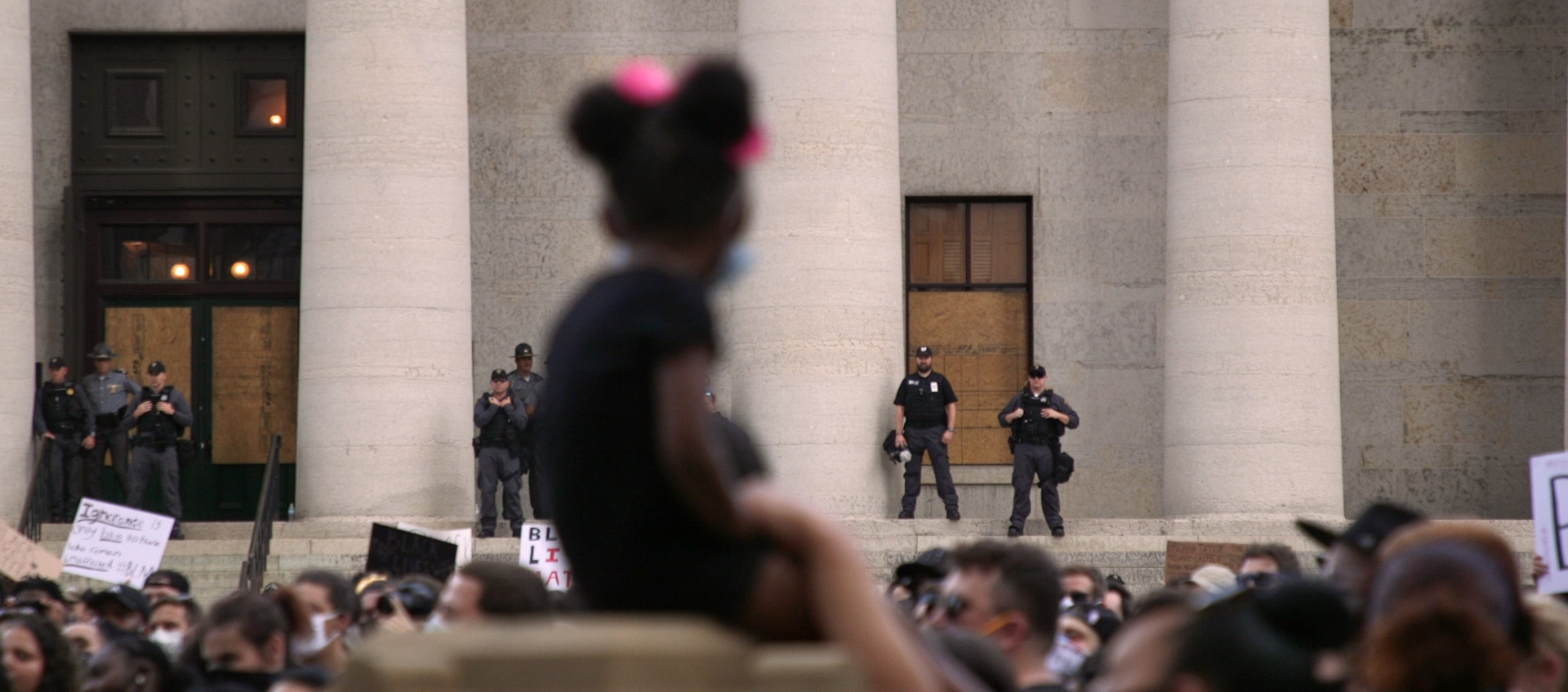 A child in the foreground sits and watches a protest. Police officers also watch.