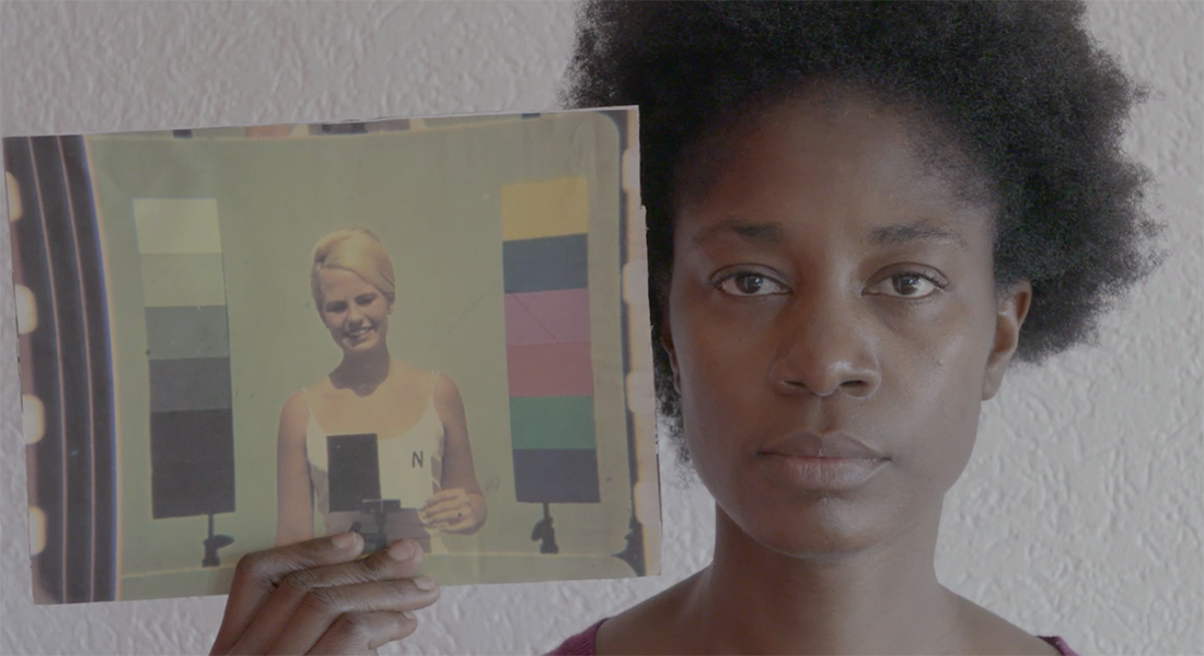 A black woman holds up an image on paper of a white woman flanked by color test patterns