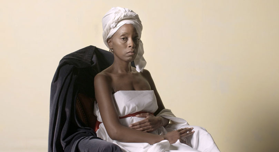 A black woman in a white dress sits on a chair draped with black velvet against a beige background