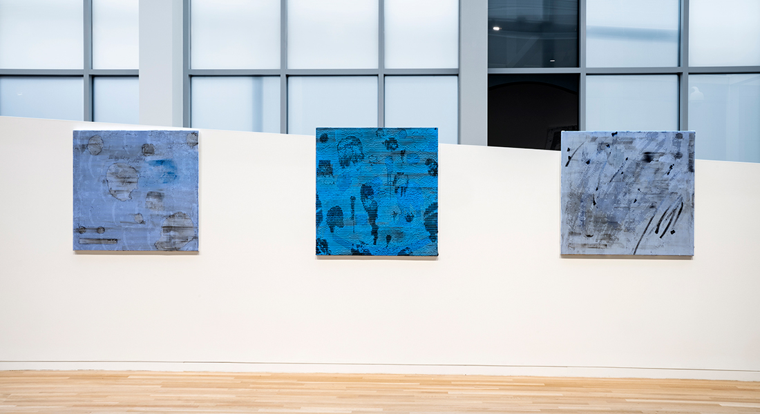 Three blue abstract canvases with gray and black markings hang on a trapezoid shaped wall in the Wexner Center galleries; a metal and glass wall is in the background and a blonde wood floor is visible in the foreground