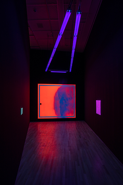 A view of three abstract paintings hung in a special wedge-shaped space in the Wexner Center galleries lit only with ultraviolet lights, visibly hanging from the ceiling. 