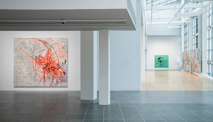 A view of three large abstract canvases hanging in the Wexner Center's galleries, with one canvas with red brushstrokes in the foreground and a green painting and multicolored painting in the background. The walls are white, the floors are wood and dark marble, and there is a glass and steel wall to the right.