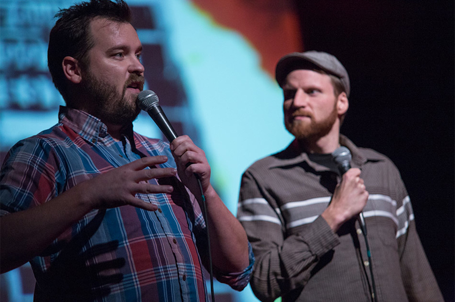 A man with a short, trimmed beard holds a microphone and speaks to an audience. next to him is a man with a neatly trimmed beard who wears a hat. He also holds a microphone
