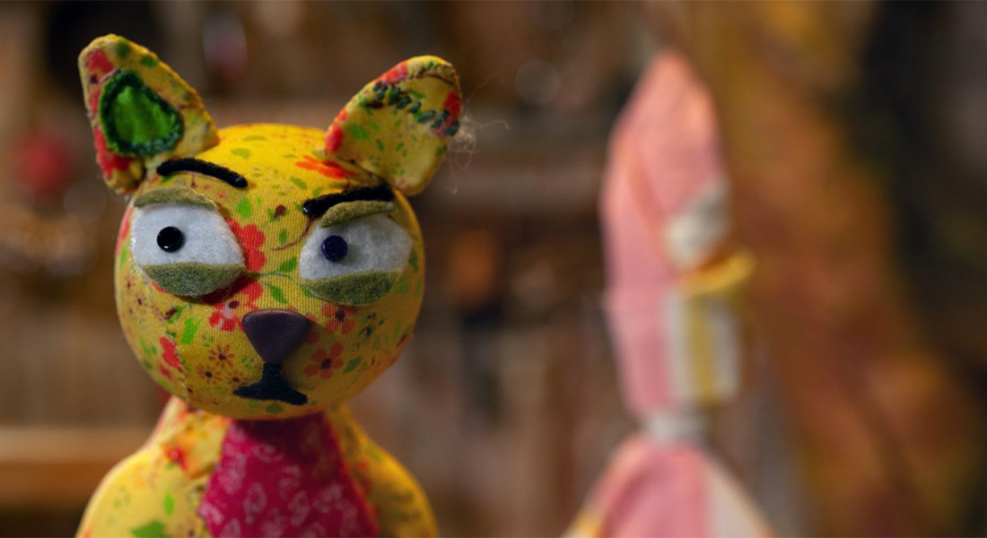 A catlike creature made of mostly yellow patterned fabric has quizzical expression in this animated still