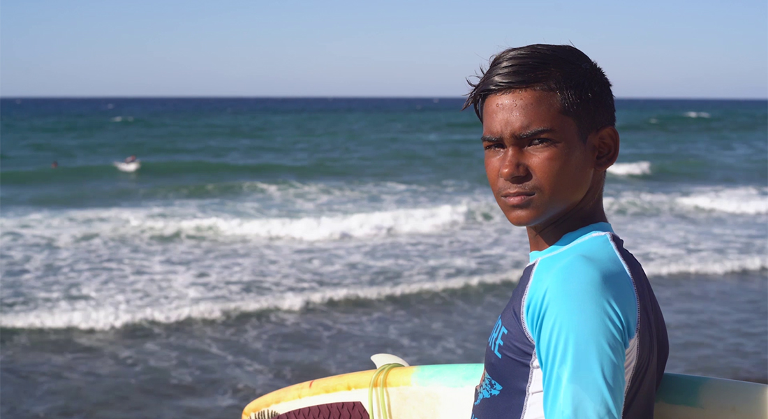 A boy with dark hair looks at the camera while holding a surfboard in front of an ocean