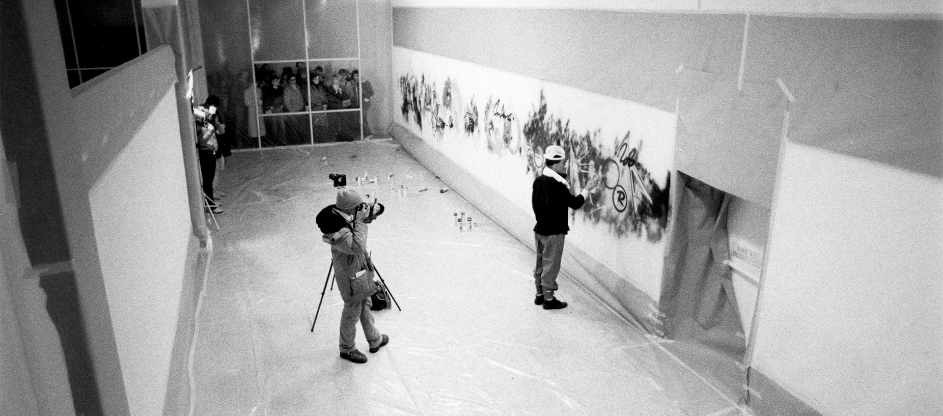 A black-and-white photo of artist Futura spray painting a long, wall-length work in a gallery lined in plastic film. Photographers film film from the center of the gallery and its doorway.