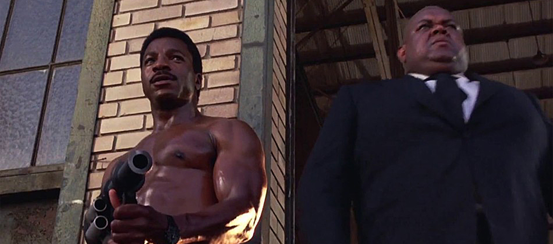Carl Weathers, muscular and shirtless, holds a grenade launcher while standing next to a bald man in a dark suit