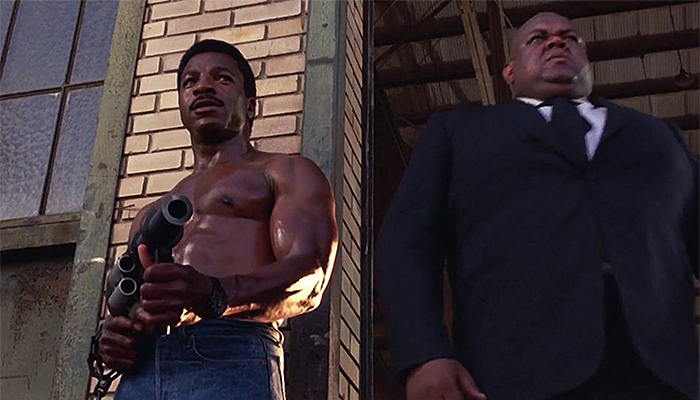 Carl Weathers, muscular and shirtless, holds a grenade launcher while standing next to a bald man in a dark suit