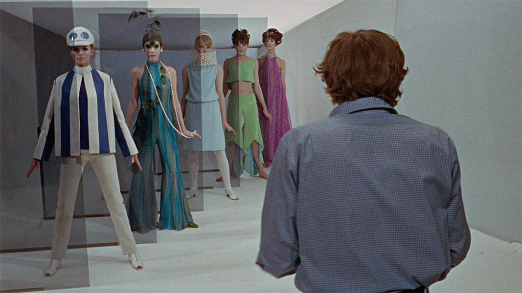 A man in a blue shirt and shaggy hair stands with his back to us. He is looking forward at a row of five fashion models in stylish, mid-1960s mod attire